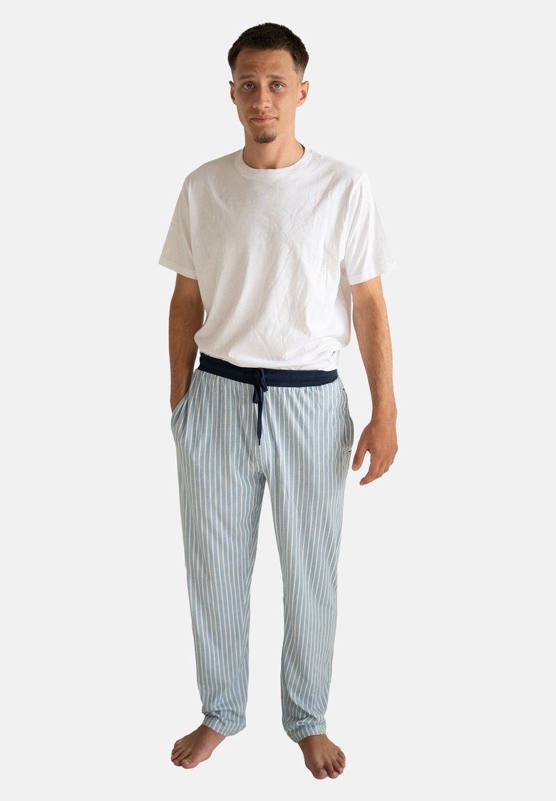 Pantalones de pijama a rayas azul claro con una cinturilla elástica azul marino y cordón; se usan con una camiseta blanca simple; el modelo está descalzo.