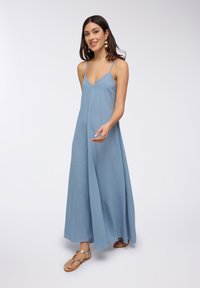 Abito maxi blu chiaro con scollatura a V, spalline sottili e una linea fluida. Il tessuto ha un aspetto strutturato. Abbinato a sandali dorati.