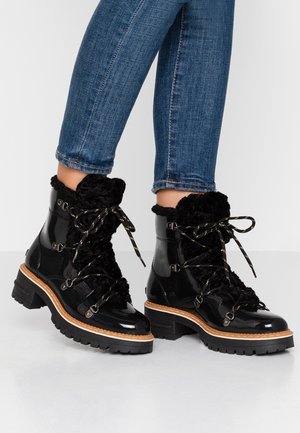 Botas de tobillo de charol negro con forro de pelo negro, cordones dorados, acentos metálicos y una suela de goma gruesa con un detalle de madera.