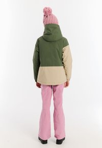 Farbenblockierte Jacke in Grün und Beige, mit Kapuze. Kombiniert mit pinkfarbenen Schlaghosen und schwarzen Stiefeln. Strickmütze in Pink mit Pompon.