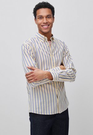 Lachende man met kort krullend haar die een crèmewit overhemd met blauwe verticale strepen draagt, met gekruiste armen, staande tegen een effen lichte achtergrond.