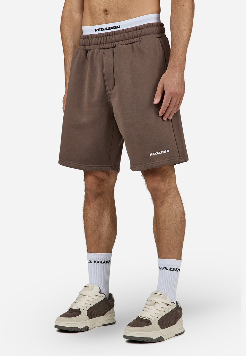 Bruna bomullsblandade shorts med elastisk midja, sidofickor och vit "PEGADOR"-logotyp. Matchas med vita strumpor och beige-bruna sneakers.