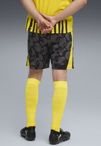 Tricou sportiv galben cu dungi verticale negre, asortat cu pantaloni şort negri cu un model texturat şi ciorapi galbeni până la genunchi, purtat cu ghete negre.