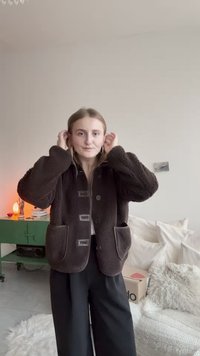 Braune Fleecejacke mit durchgehendem Knopfverschluss, großen Taschen und strukturierter Oberfläche, kombiniert mit schwarzen, weit geschnittenen Hosen.