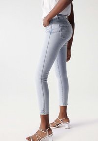 Jeans de tiro alto en azul claro con un ajuste slim, que presentan un bolsillo trasero desgastado, combinados con tacones blancos con tiras. Textura de mezclilla suave.