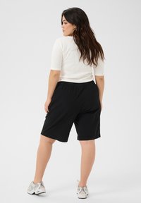 Shorts noirs arrivant au genou avec une ceinture élastique, associés à un haut blanc ajusté. Le mannequin a les cheveux longs et porte des chaussures de sport légères.