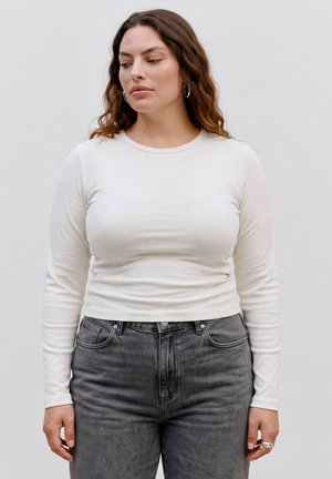 Kvinde med langt brunt hår iført hvid langærmet skjorte, grå jeans med høj talje, sølv creol-øreringe og ring, står mod en ensfarvet lys baggrund.