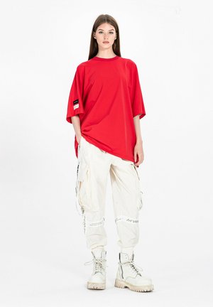 Joven mujer de pie, vistiendo una camiseta roja holgada, pantalones cargo blancos con detalles de texto y botas blancas con cordones sobre un fondo blanco liso.