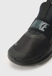 Chaussure de sport noire avec une tige en mesh texturé et une semelle en caoutchouc. Présente un logo Nike gris bien visible sur une sangle élastique. Design à bout arrondi.
