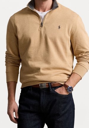 Man draagt een beige quarterzip trui met polo-logo, wit ondershirt, donkere spijkerbroek, bruine riem en een bruin leren horloge met blauwe wijzerplaat.
