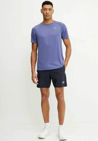 Maglietta sportiva viola a maniche corte con logo, abbinata a pantaloni corti neri con dettagli bianchi, completata da sneakers bianche.