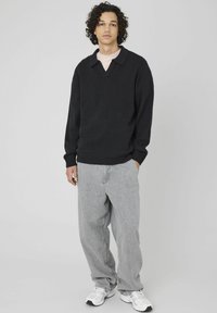 Un jeune homme avec des cheveux bouclés portant un pull noir sur une chemise rose clair, un jean gris ample et des baskets blanches, se tenant avec une main dans la poche.