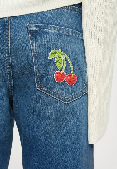 Blauwe denim jeans zak met geborduurde rode kersen en groene bladeren, in combinatie met een witte gebreide sweater mouw.