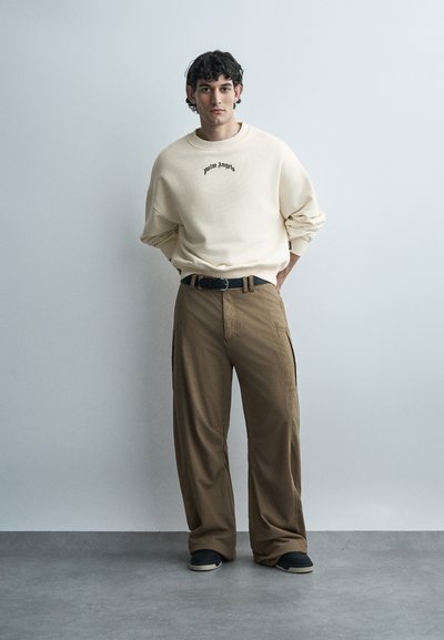 Νεαρός άνδρας με σγουρά μαλλιά στέκεται μπροστά σε έναν απλό τοίχο, φορώντας εκρού φούτερ, καφέ φαρδύ παντελόνι, μαύρη ζώνη και μαύρα παπούτσια.