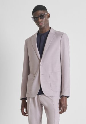 Man in een lichtbeige pak, donkere blouse eronder en grote donkere zonnebril, staand tegen een effen lichte achtergrond.