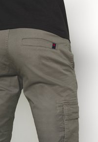 Zelene cargo hlače s glatkom teksturom, koje imaju stražnji džep s mornarsko plavim i crvenim detaljima. Hlače su ravnog kroja.