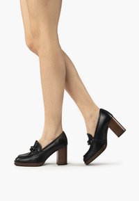 Chaussures à plateforme en cuir noir avec un bout rond, un détail en nœud et un talon en bois empilé. Texture lisse et design structuré.