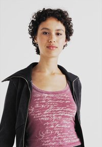 Black zippered jacket over a pink tank top featuring white script text. The tank has a rounded neckline and a relaxed fit.