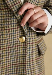 Vlněný blejzer v houndstooth vzoru v zelených, zlatých a červených odstínech, s výrazným zlatým knoflíkem a viditelnými bílými manžetami košile.