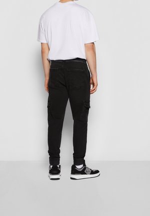 Man draagt zwarte cargobroek, wit T-shirt, witte sokken en zwart-witte sneakers, staat met het gezicht naar een grijze muur gericht.