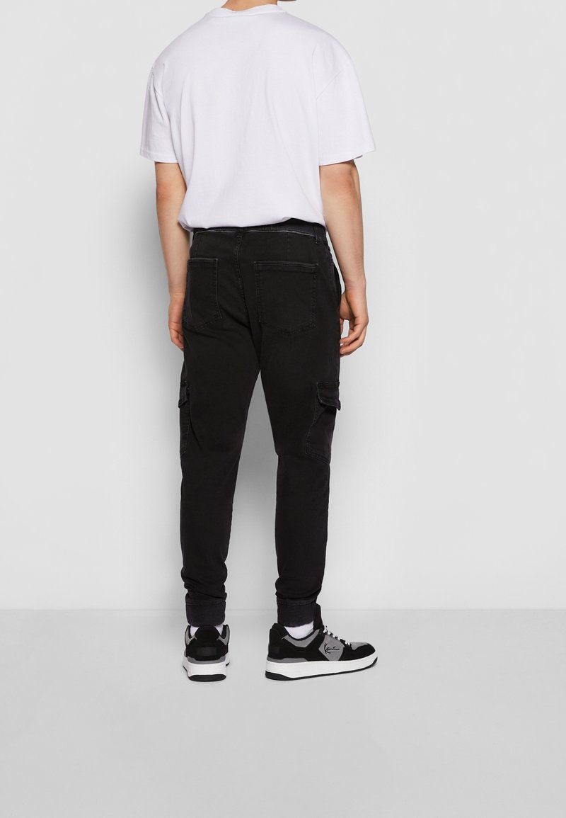 Homme portant un pantalon cargo noir, un t-shirt blanc, des chaussettes blanches et des baskets noires et blanches, se tenant face à un mur gris.
