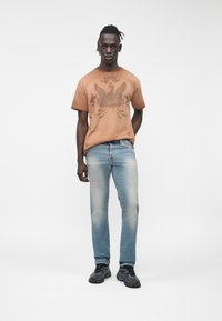 Camiseta gráfica marrón con diseño de águila, combinada con jeans azul claro descoloridos y zapatos deportivos negros. De pie contra un fondo liso.