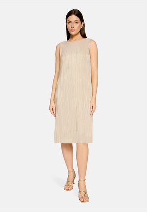 Femme debout portant une robe beige plissée sans manches longueur genou et des sandales à talons dorées à brides sur fond uni.