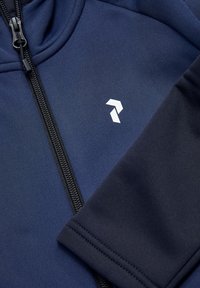 Marineblå zip-jakke med tekstureret stof, sort lynlås og et hvidt geometrisk logo på øverste venstre side.