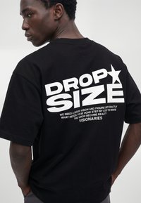 Giovane uomo che indossa una T-shirt nera con grande testo bianco "DROP SIZE" e testo più piccolo sulla visione e i visionari sul retro.