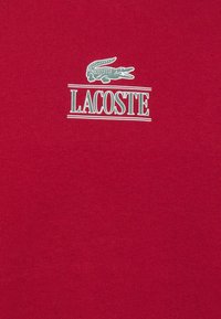 Červené bavlnené tričko s zeleným logom krokodíla a textom "LACOSTE" v bielej farbe, tlačeným na strednej časti hrudi. Mäkka, hladká textúra.
