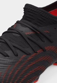 Μαύρο αθλητικό παπούτσι με υφή στο πάνω μέρος, κόκκινες ραφές που μοιάζουν με σχέδια, μαύρα κορδόνια και κόκκινη εσωτερική επένδυση ορατή.