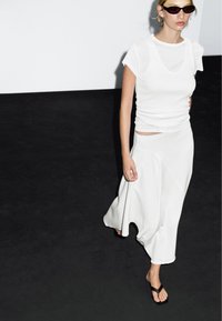 Woman walking on black floor wearing white sleeveless top, long white skirt, black sunglasses, and black sandals.