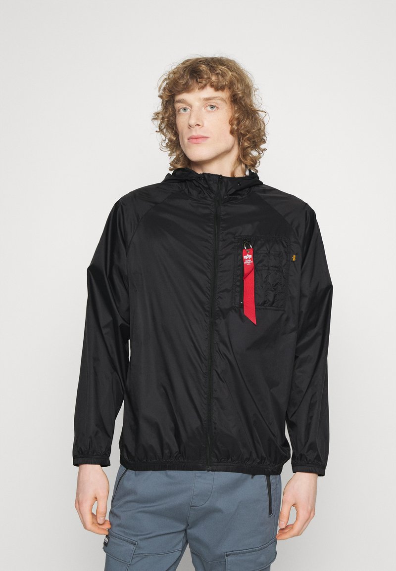 Schwarze, leichte Jacke mit Kapuze, Reißverschluss vorne, elastischen Bündchen und einer Brusttasche mit einem roten Akzent-Tag. Glattes, glänzendes Material.