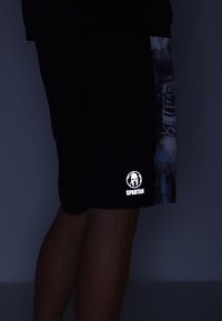 Homme portant un short noir avec un panneau latéral gris à motifs et un logo de casque spartiate blanc près de l'ourlet.