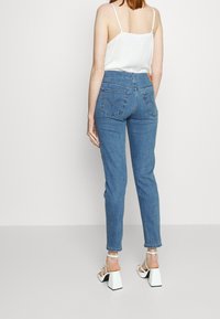 Femme portant un jean taille haute bleu, un top blanc sans manches et des sandales blanches à talons carrés, debout devant un fond clair uni.