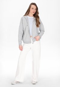 Šedá mikina na zip s volným střihem, která je kombinována s pruhovaným tričkem a volnými bílými kalhotami. Oblečení vytváří ležérní, vrstvený vzhled.