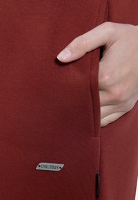 Maronfarbenes, eng anliegendes Kleidungsstück mit gerippter Textur. Verfügt über eine Seitentasche und ein kleines metallisches Logo-Tag mit der Aufschrift "SMILODOX" in der Nähe des Saums.