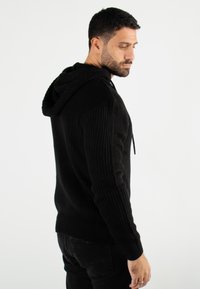 Sweat à capuche noir en maille avec un motif texturé, doté d'un cordon de serrage, de poignets et d'un bas côtelés. Porté avec un pantalon noir, vu de profil.