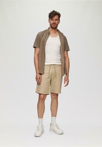 Chemise courte à manches courtes marron clair sur un débardeur blanc, accompagnée de shorts beige et de baskets blanches avec des accents gris clair.