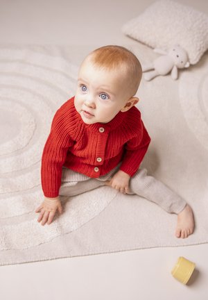 Roter Strickcardigan mit strukturiertem Muster und Holzknöpfen, kombiniert mit grauen Strickhosen. Gelber Spielzeugbecher neben einem runden, strukturierten Teppich.