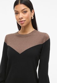 Pulover s chevron vzorom v rjavi in črni barvi, okroglim izrezom, dolgimi rokavi in mehkimi vlakni. Kontrastne barvne blokade poudarjajo obliko.