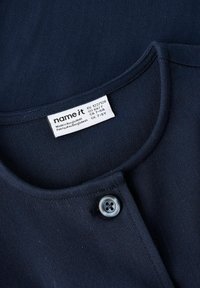 Temno modra tkanina z nežno teksturo, z zaobljenim izrezom in sivim gumbom spredaj. Pripeta etiketa za nego.