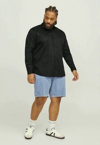 Uomo con capelli intrecciati, che indossa una camicia nera con bottoni, pantaloncini di jeans azzurri chiari, calze bianche e sneakers bianche, in piedi contro uno sfondo neutro.