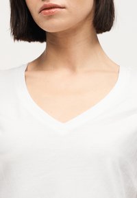 Γυναίκα με κοντά σκούρα μαλλιά που φοράει λευκό πουκάμισο με V-λαιμόκοψη, εμφανίζοντας το λαιμό και το πάνω μέρος του στήθους σε ουδέτερο φόντο.