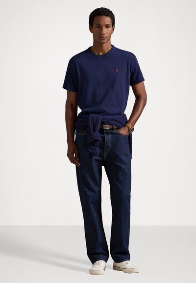 Tengerész navy crew neck póló, pamutból, kis piros logóval. Sötét farmernadrág, laza fazon, és fehér sportcipő. Kardigán megkötve a derékon.