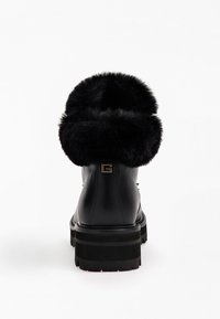 Svart läderkänga med tjock sula och vikta svarta pälskanter, sedd bakifrån mot en vit bakgrund.