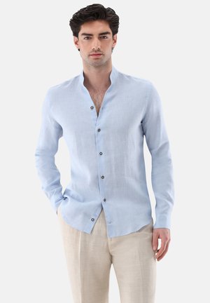 Mann trägt hellblaues Hemd mit Mandarinkragen und beige Hose, steht mit einer Hand in der Tasche, neutraler Hintergrund.