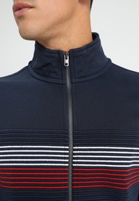 Giacca a zip blu navy con colletto alto, caratterizzata da strisce orizzontali bianche e rosse. Il tessuto è strutturato e liscio.