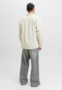 Man die met zijn rug naar de camera staat, gekleed in een crèmekleurig corduroy shirt en losse, vergrijsde blauwe jeans tegen een vlakke witte achtergrond.