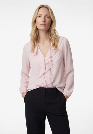 Blondynka w jasnoróżowej bluzce z falbanami i długimi rękawami oraz czarnych, dopasowanych spodniach, ręce w kieszeniach, neutralny wyraz twarzy.